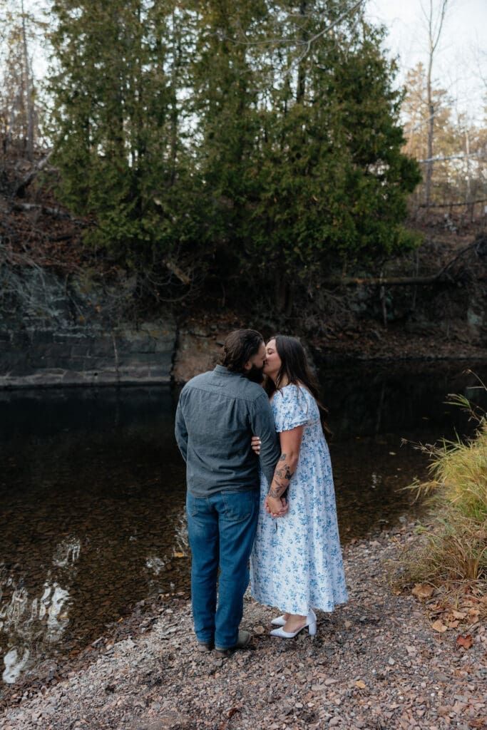 Engaged couple sharing a kiss near a wooded area and water at Glensheen Mansion in Duluth, Minnesota.