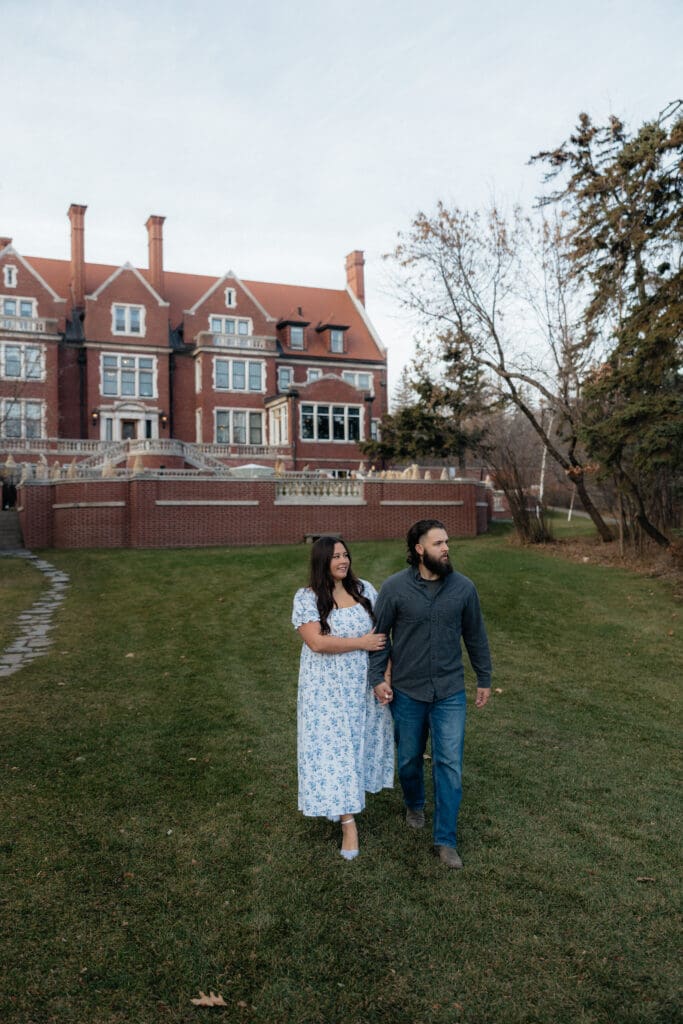 Engaged couple walking hand in hand across the lawn of Glensheen Mansion in Duluth, Minnesota, during their engagement session.