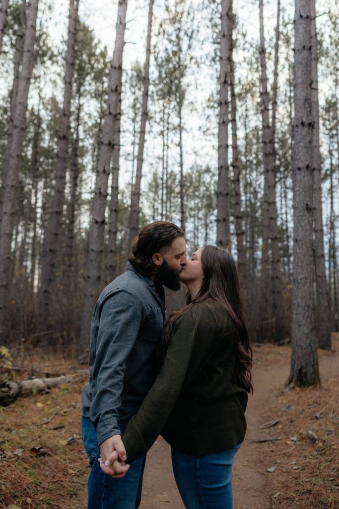 Engaged couple kissing in a serene pine forest, holding hands during their adventure engagement session.