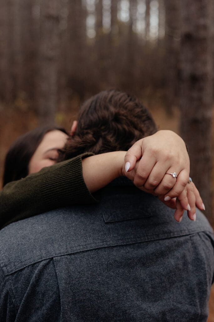 Close-up of an engagement ring as a woman embraces her fiancé in a cozy forest setting.