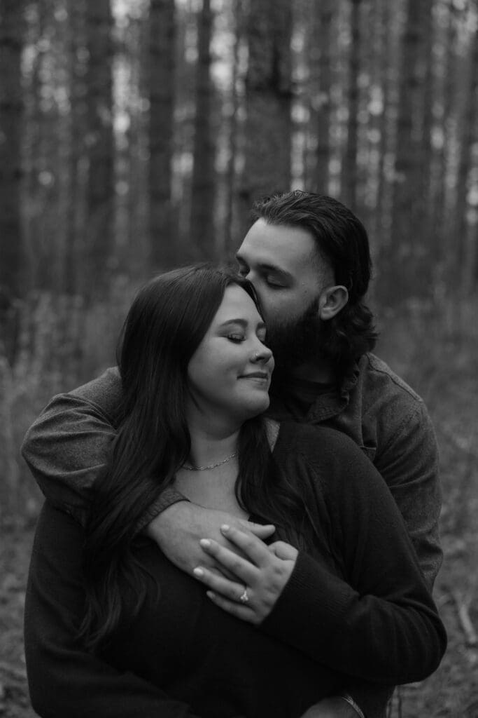 Black-and-white engagement portrait of a woman smiling while her fiancé wraps his arms around her.