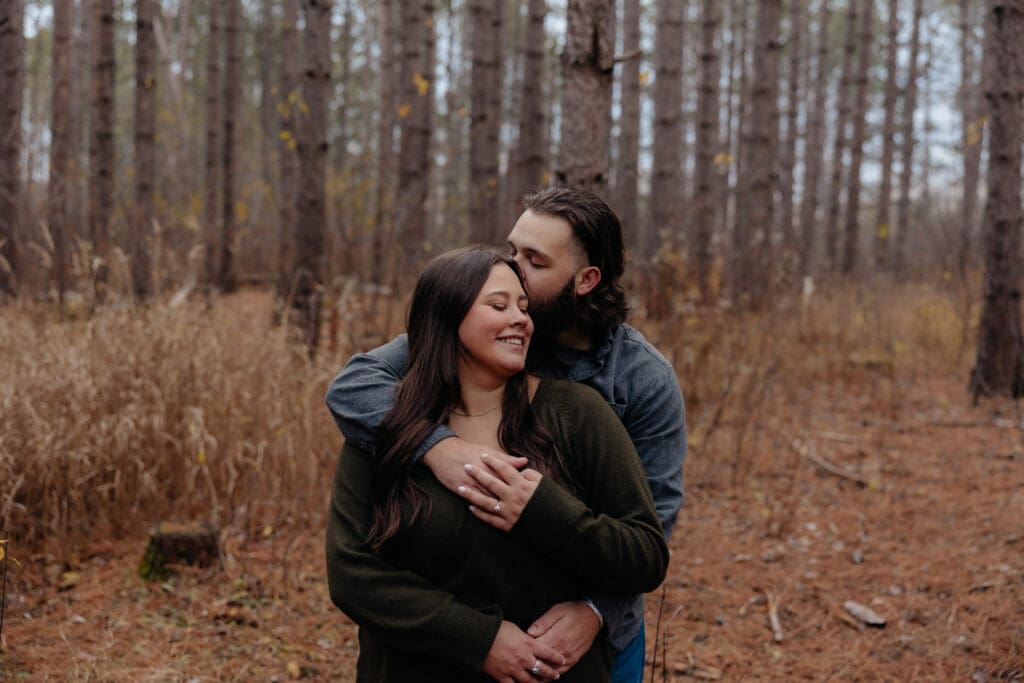 Man embracing his fiancée from behind, whispering into her ear in a wooded setting near Duluth.