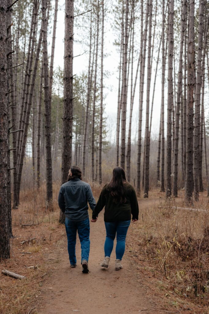 Engaged couple walking hand in hand down a scenic pine-lined path in Duluth, Minnesota.